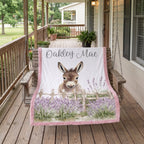 A personalized blanket draped over a wooden porch swing on a rustic wooden porch. The blanket features a white background with a printed illustration of a donkey behind a wooden fence in a field of purple lavender.