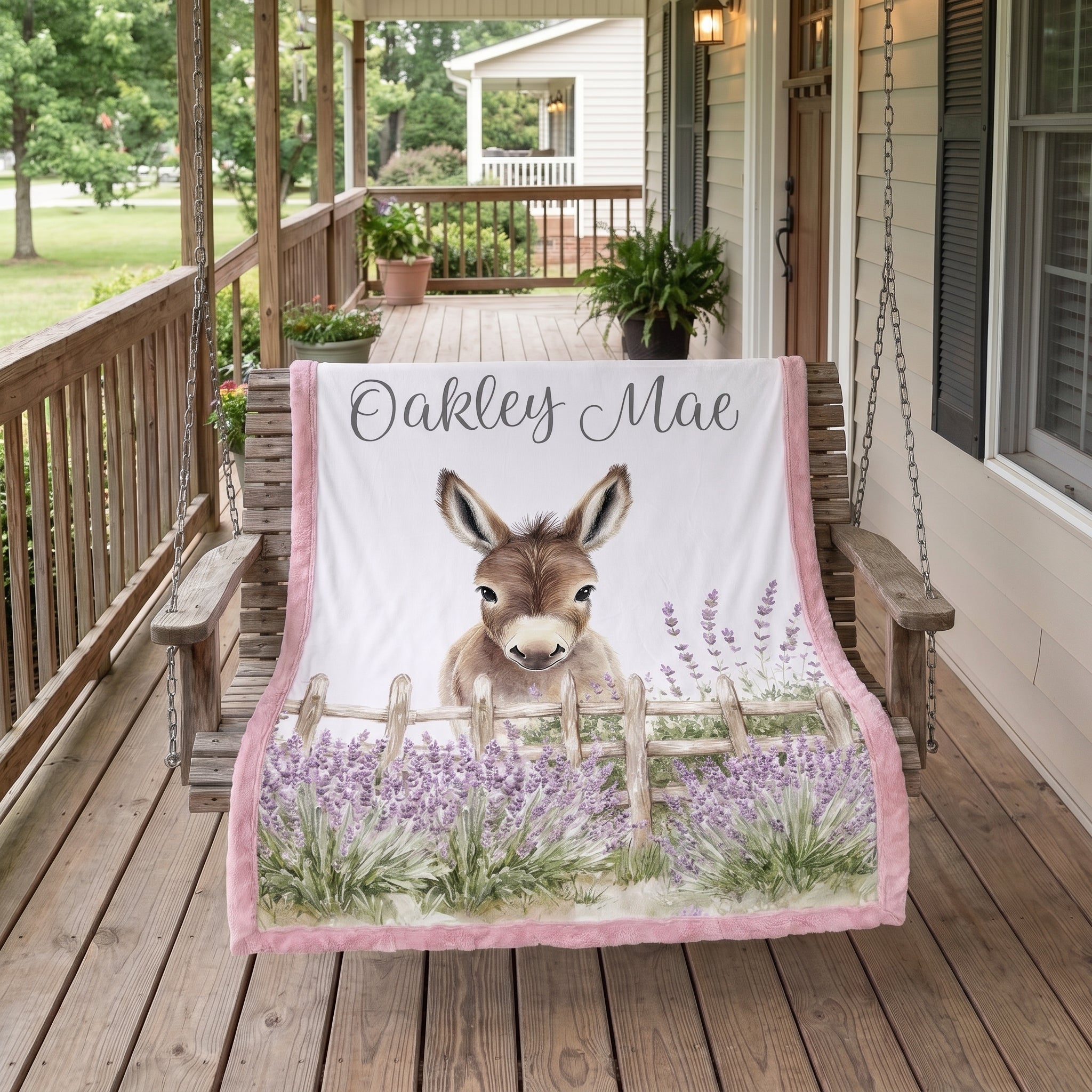 A personalized blanket draped over a wooden porch swing on a rustic wooden porch. The blanket features a white background with a printed illustration of a donkey behind a wooden fence in a field of purple lavender.