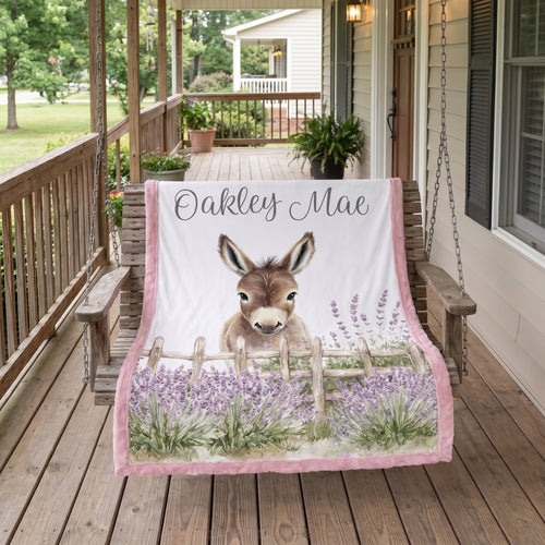 A personalized blanket draped over a wooden porch swing on a rustic wooden porch. The blanket features a white background with a printed illustration of a donkey behind a wooden fence in a field of purple lavender.