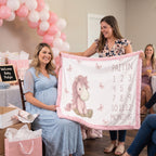 A woman in a light blue maternity dress and another woman hold up a personalized baby blanket with a pink unicorn, butterflies, and the name 'PAITYN' with birth months. The scene is set in a decorated baby shower environment with pink and white balloon arches, gift bags, and a welcome sign.