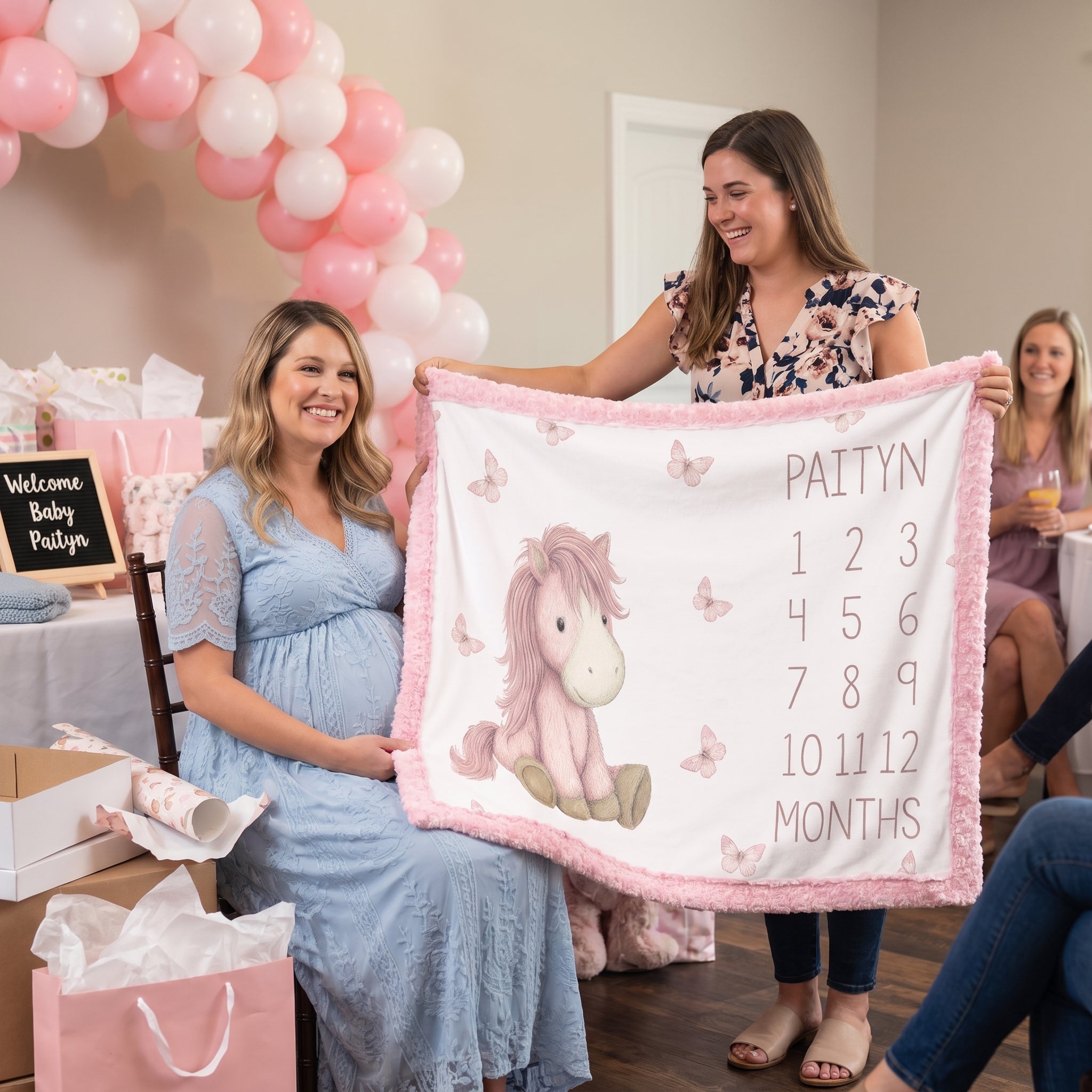 A woman in a light blue maternity dress and another woman hold up a personalized baby blanket with a pink unicorn, butterflies, and the name 'PAITYN' with birth months. The scene is set in a decorated baby shower environment with pink and white balloon arches, gift bags, and a welcome sign.