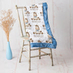 A personalized baby blanket with a white background, blue ruffled trim, and a cartoon bear pattern with the name 'BENTLEY' printed repeatedly, draped over a rustic wooden chair next to a light blue vase with dried grass.