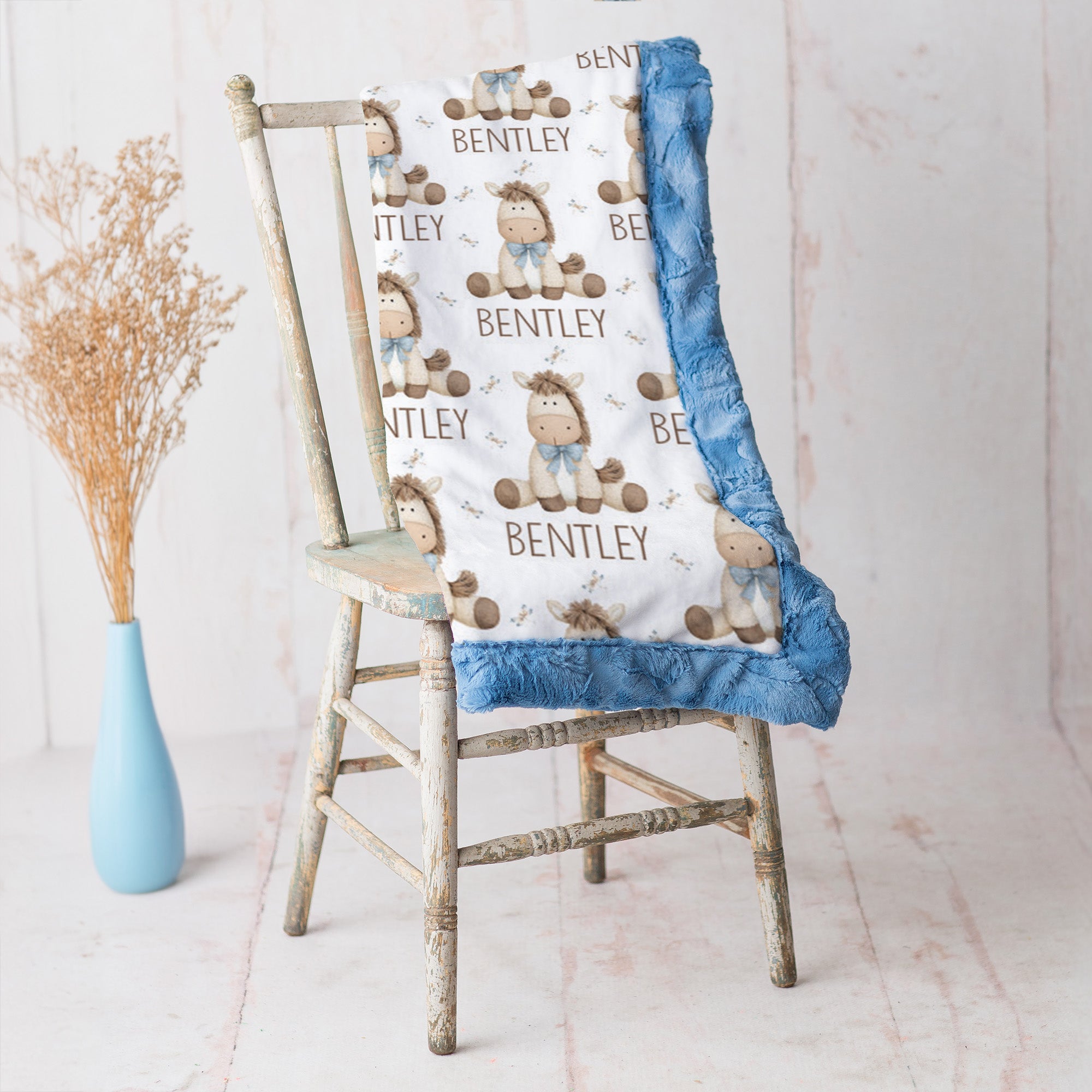 A personalized baby blanket with a white background, blue ruffled trim, and a cartoon bear pattern with the name 'BENTLEY' printed repeatedly, draped over a rustic wooden chair next to a light blue vase with dried grass.
