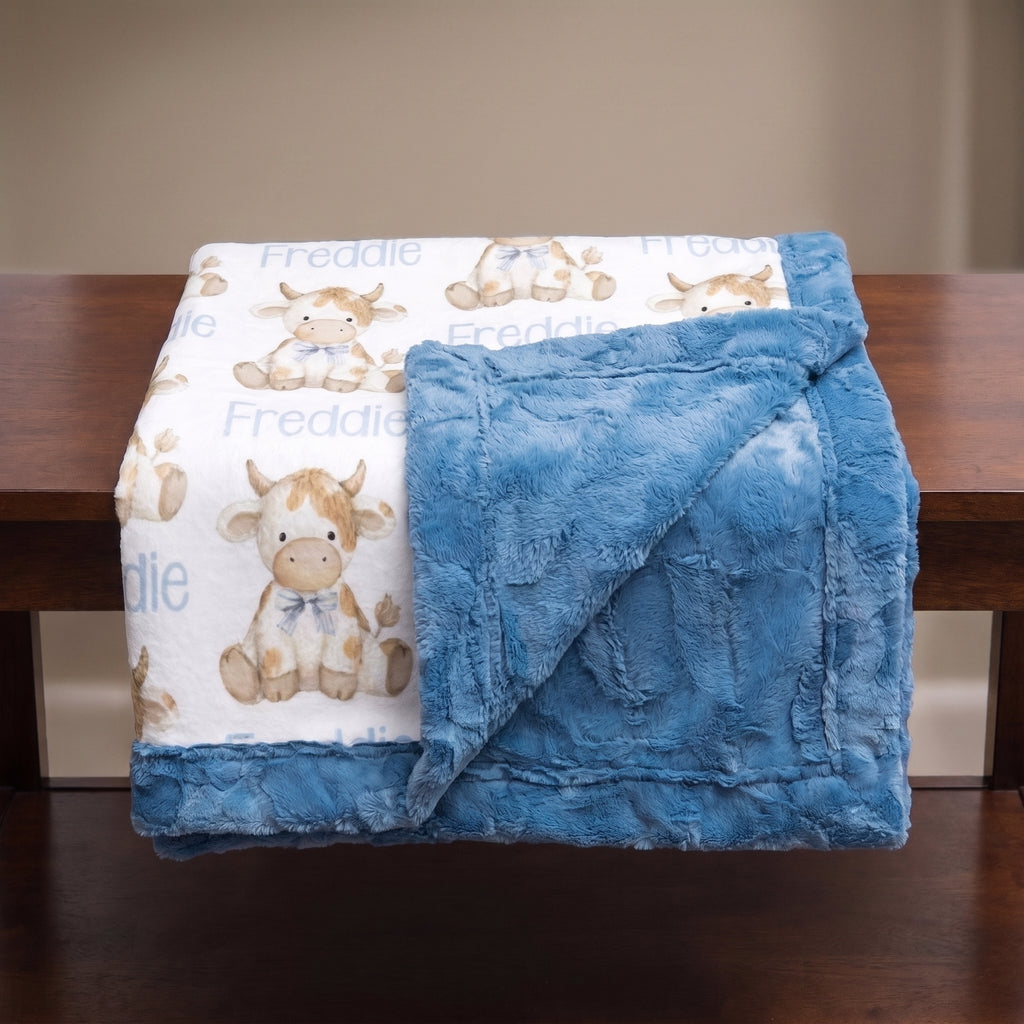 A folded baby blanket draped over a wooden table, showing a white front side with a repeating cow print and the name 'Freddie' in blue, plus a soft blue plush reverse side.