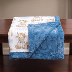 A folded baby blanket draped over a wooden table, showing a white front side with a repeating cow print and the name 'Freddie' in blue, plus a soft blue plush reverse side.