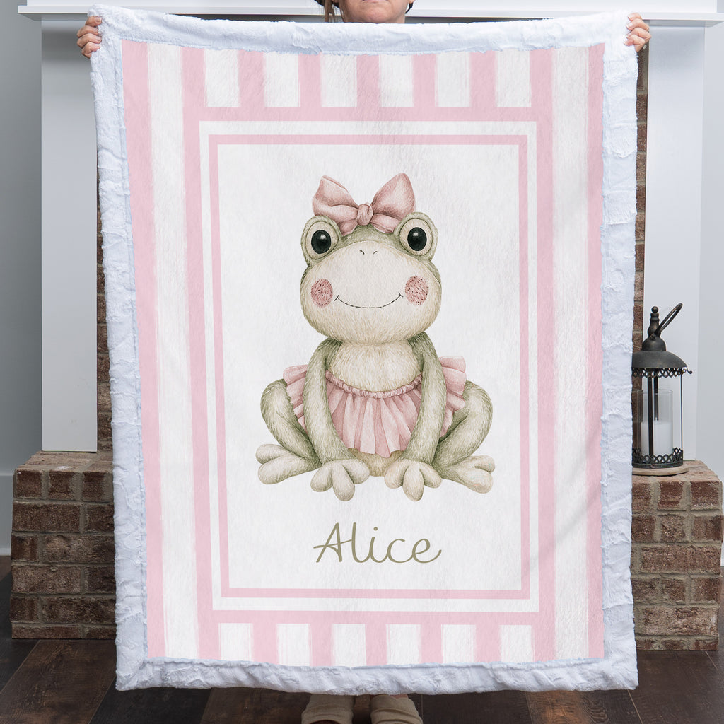 Personalized minky ballerina frog baby blanket with pink stripe border held up by fireplace