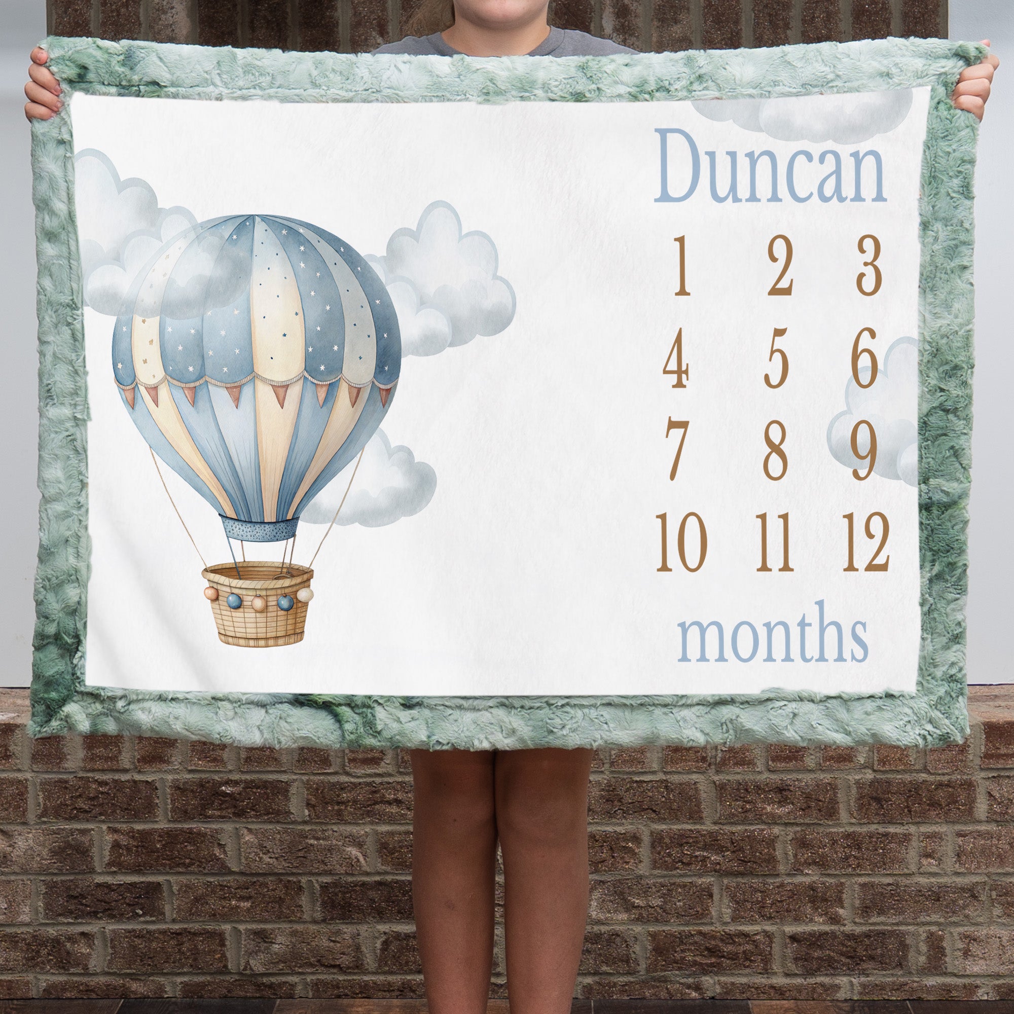Personalized hot air balloon milestone minky blanket with sage trim held up outdoors