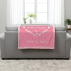 Pink blanket with text on a gray couch in a room with plants and windows.