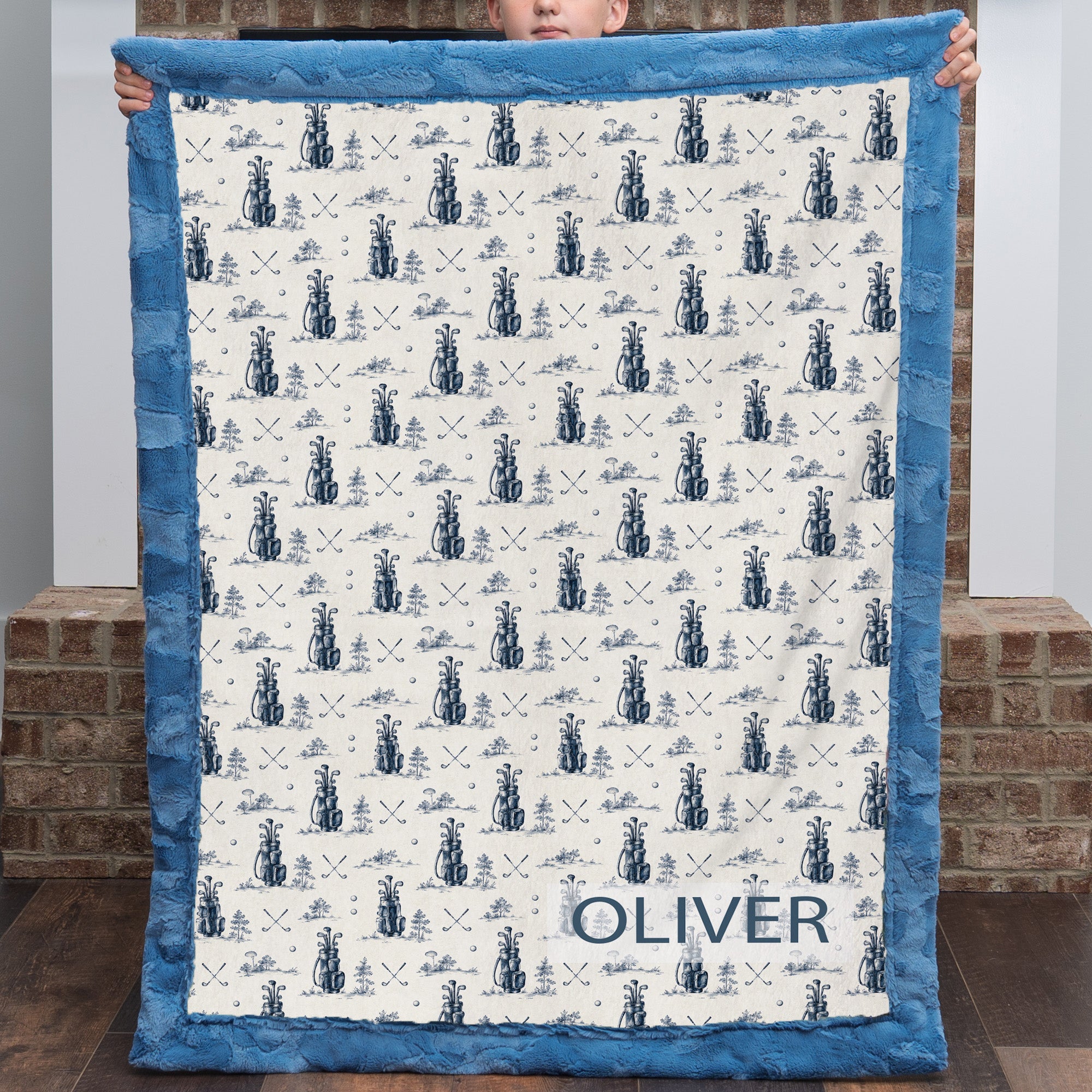 Child holding personalized golf-print minky blanket with name Oliver, blue trim, by fireplace