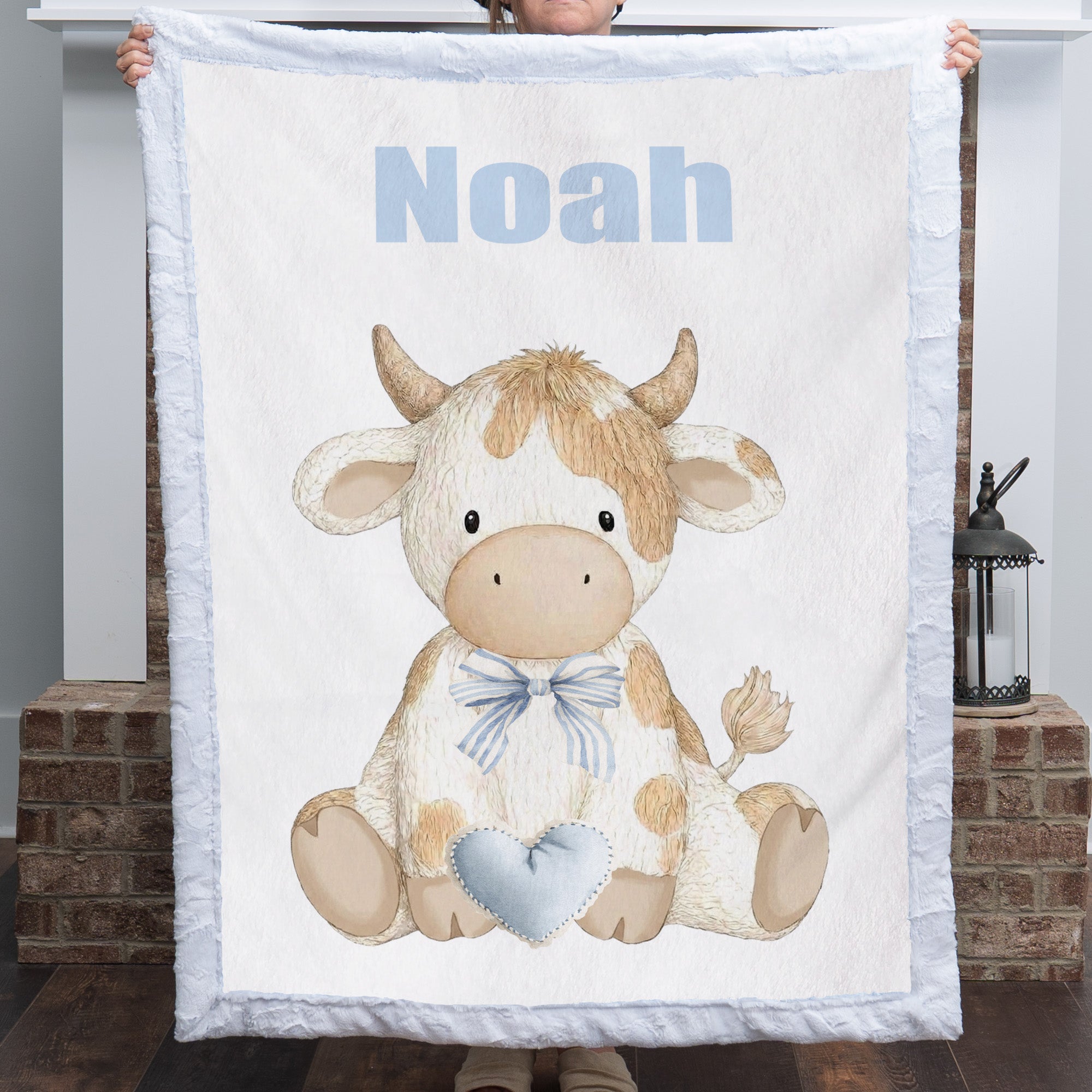 Personalized Highland cow minky baby blanket with Noah's name held up in front of brick fireplace