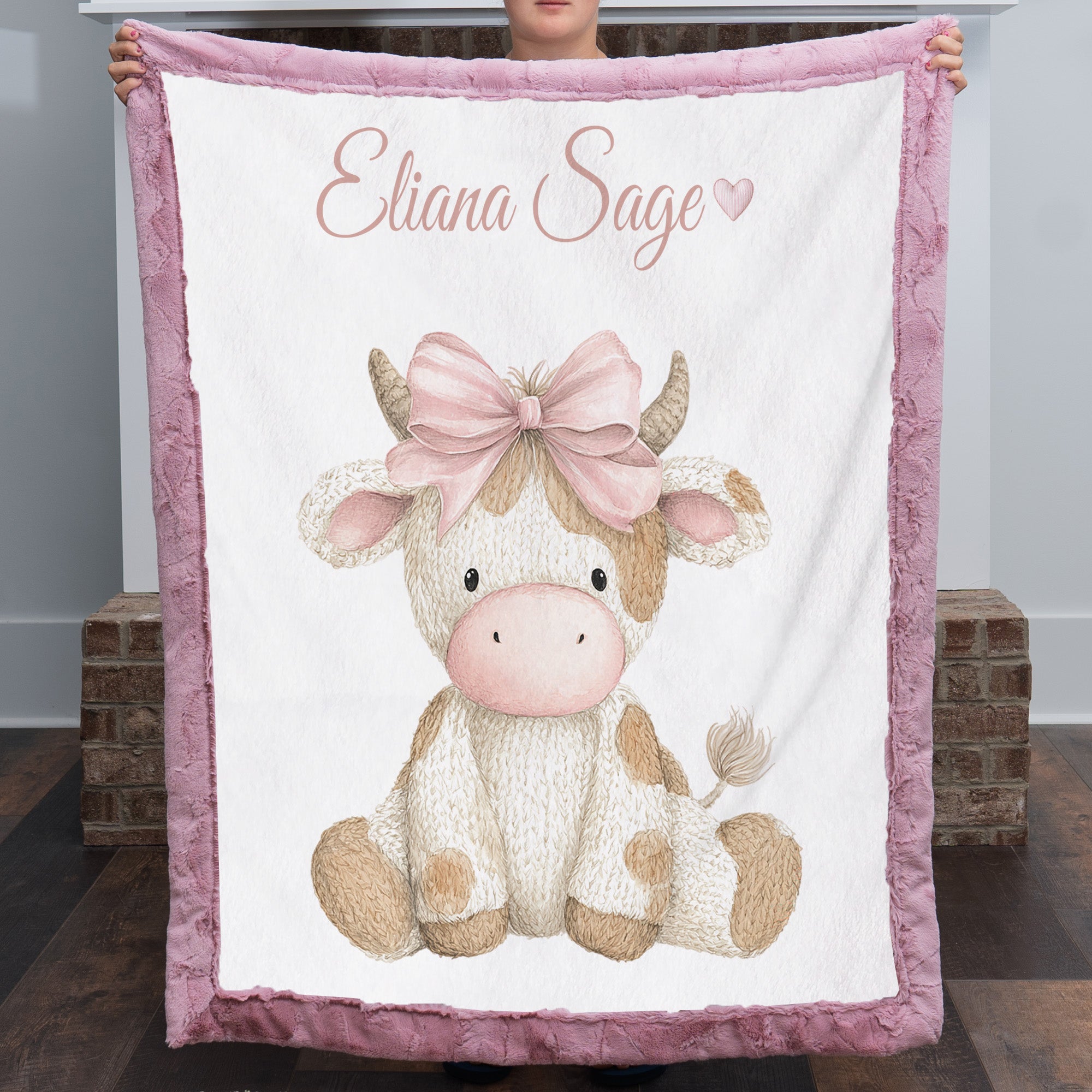 Woman holding personalized highland cow minky blanket with pink trim over fireplace mantel