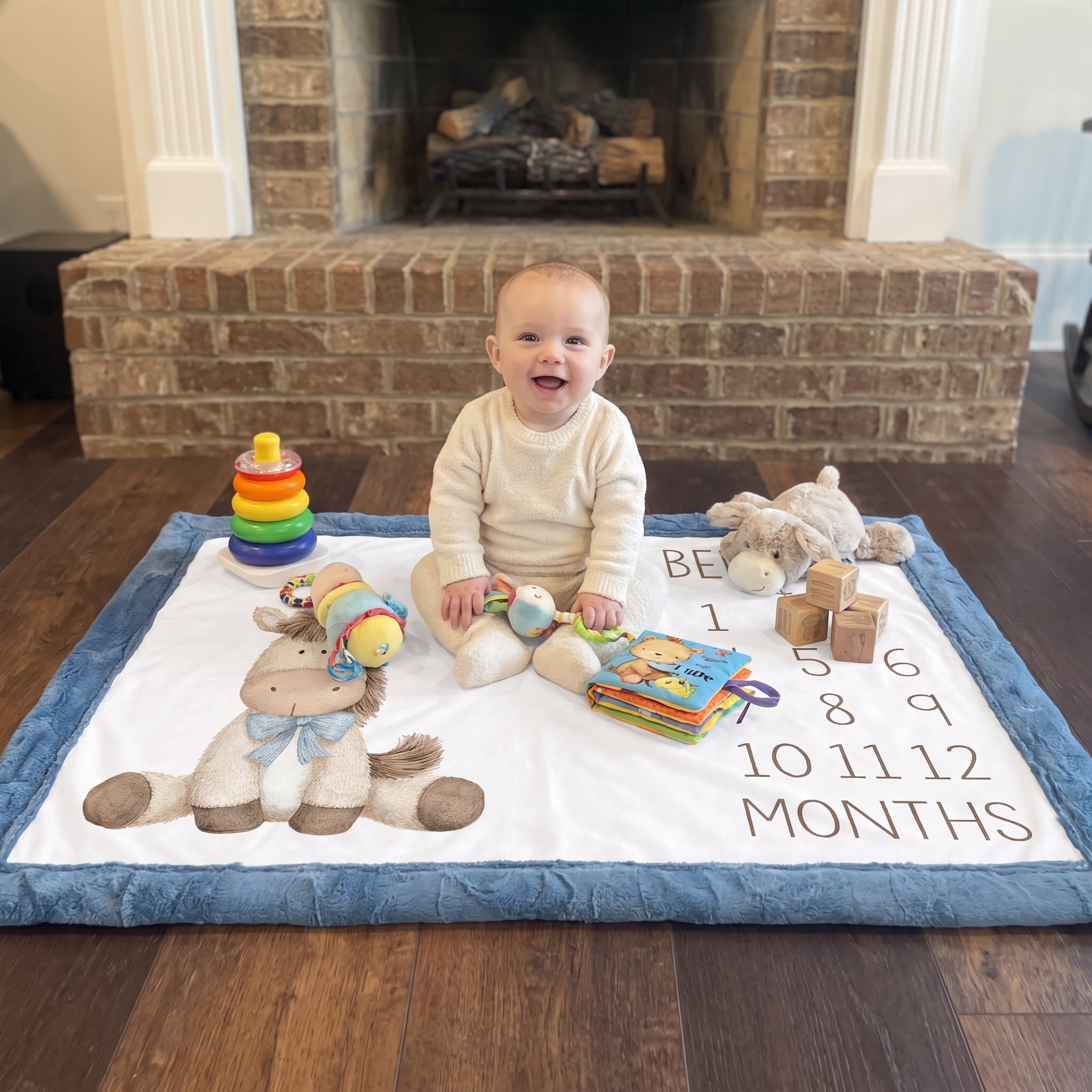 Baby sitting on personalized horse milestone blanket with toys by fireplace