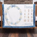 Personalized milestone baby blanket with hydrangea wreath and blue trim hanging on crib rail