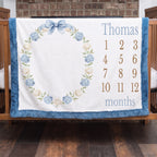 Personalized milestone baby blanket with hydrangea wreath and blue trim hanging on crib rail