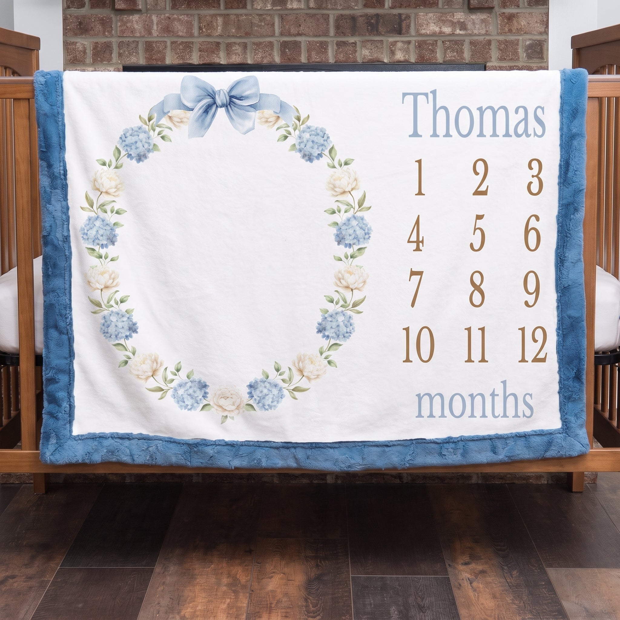 Personalized milestone baby blanket with hydrangea wreath and blue trim hanging on crib rail