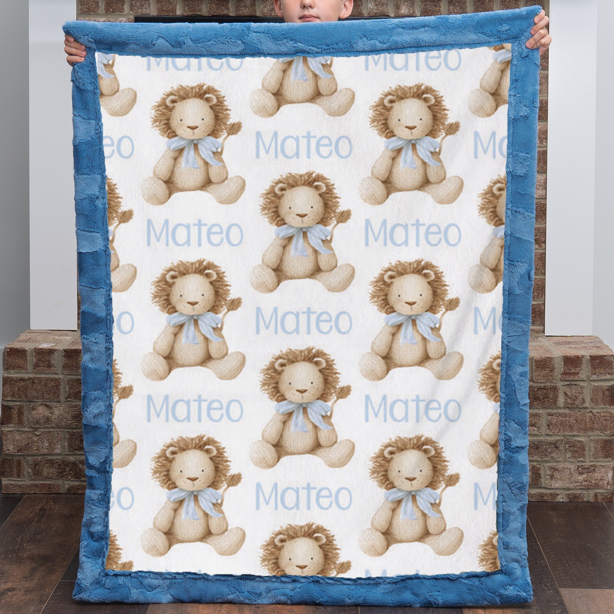 A baby blanket held up by a person, featuring a repeating pattern of teddy bears with blue bows and the name 'Mateo' printed in light blue, finished with a plush blue border.