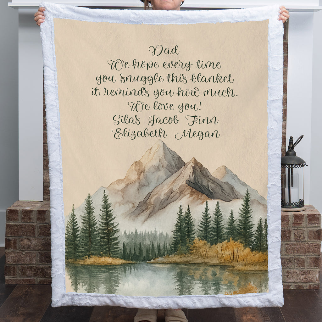 Personalized mountain lake blanket with a Dad message held up in front of a brick fireplace indoors