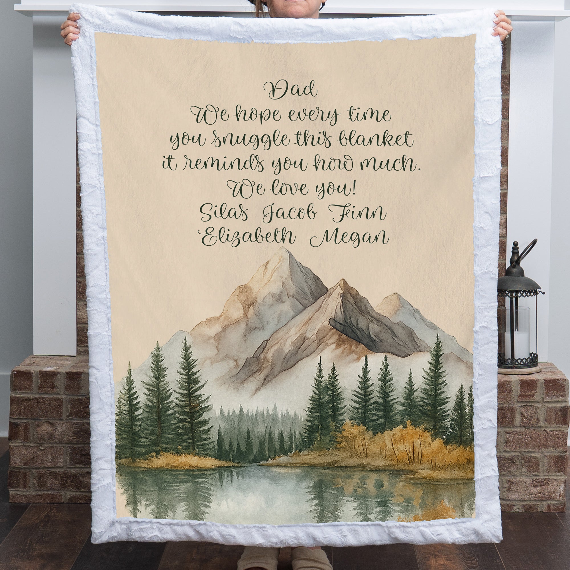 Personalized mountain lake blanket with a Dad message held up in front of a brick fireplace indoors