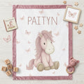 Personalized pony baby blanket with pink minky trim and butterflies, flat-laid with nursery props