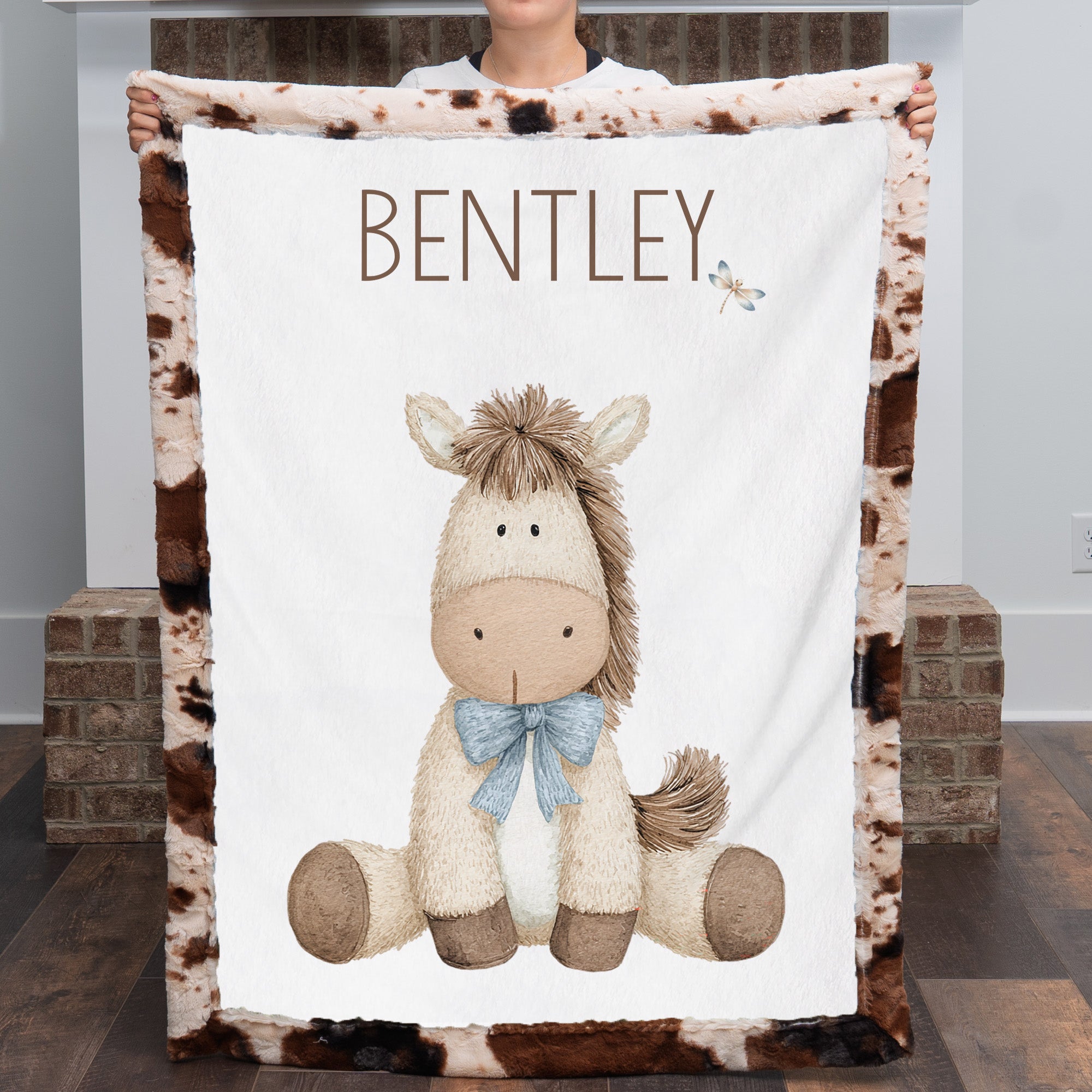 Woman holding personalized pony baby blanket with cow-print minky trim by fireplace