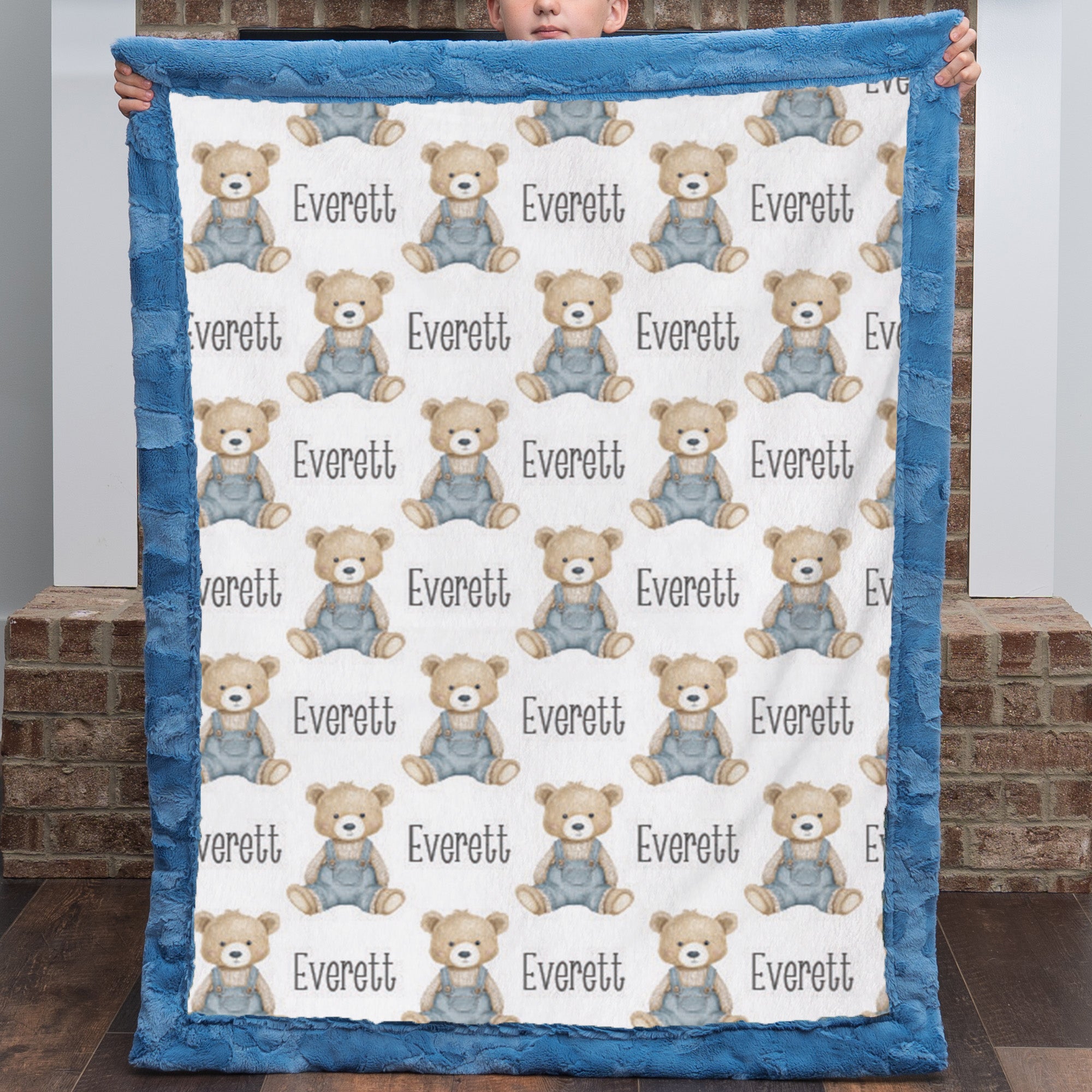 Personalized teddy bear minky baby blanket with blue fur border held up by brick fireplace
