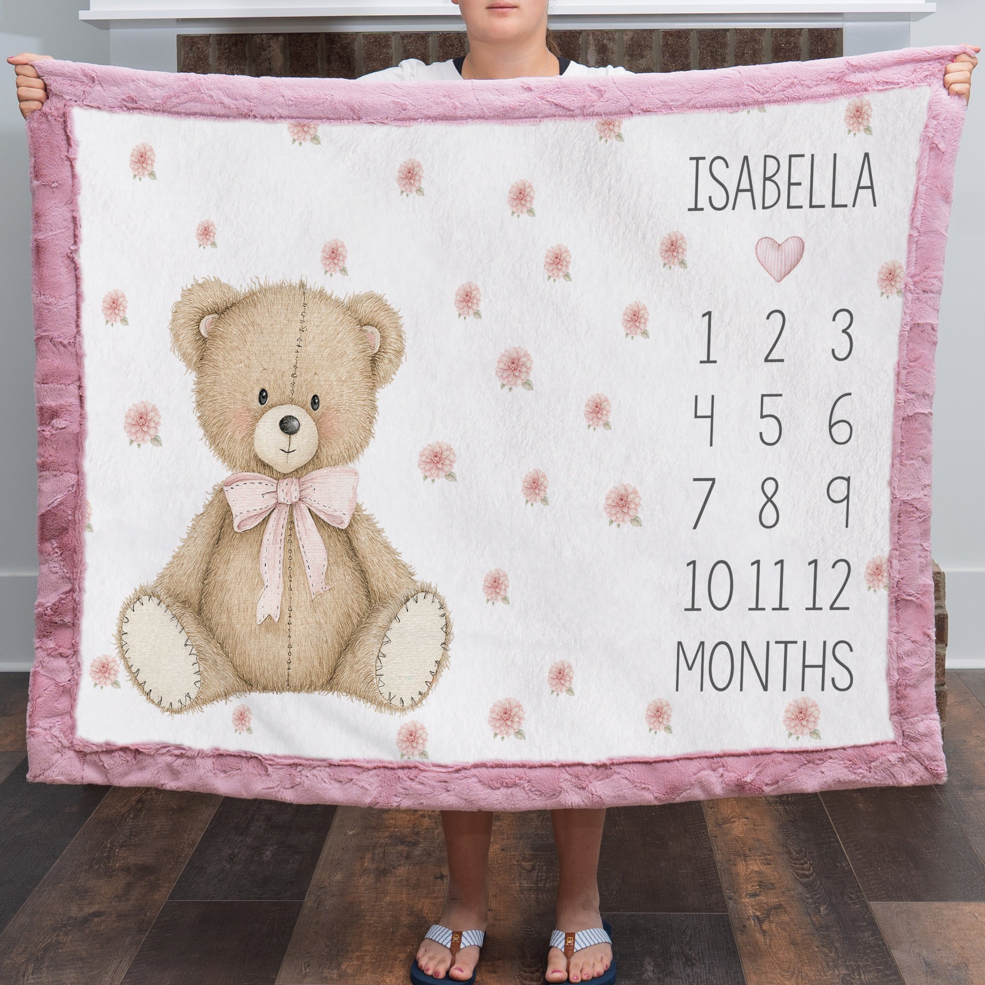 Woman holding personalized teddy bear milestone blanket with pink minky trim by fireplace