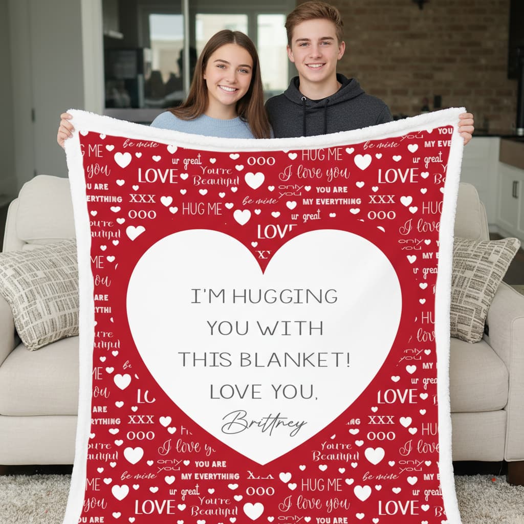 Two people holding a red blanket with heart patterns and text in a living room.