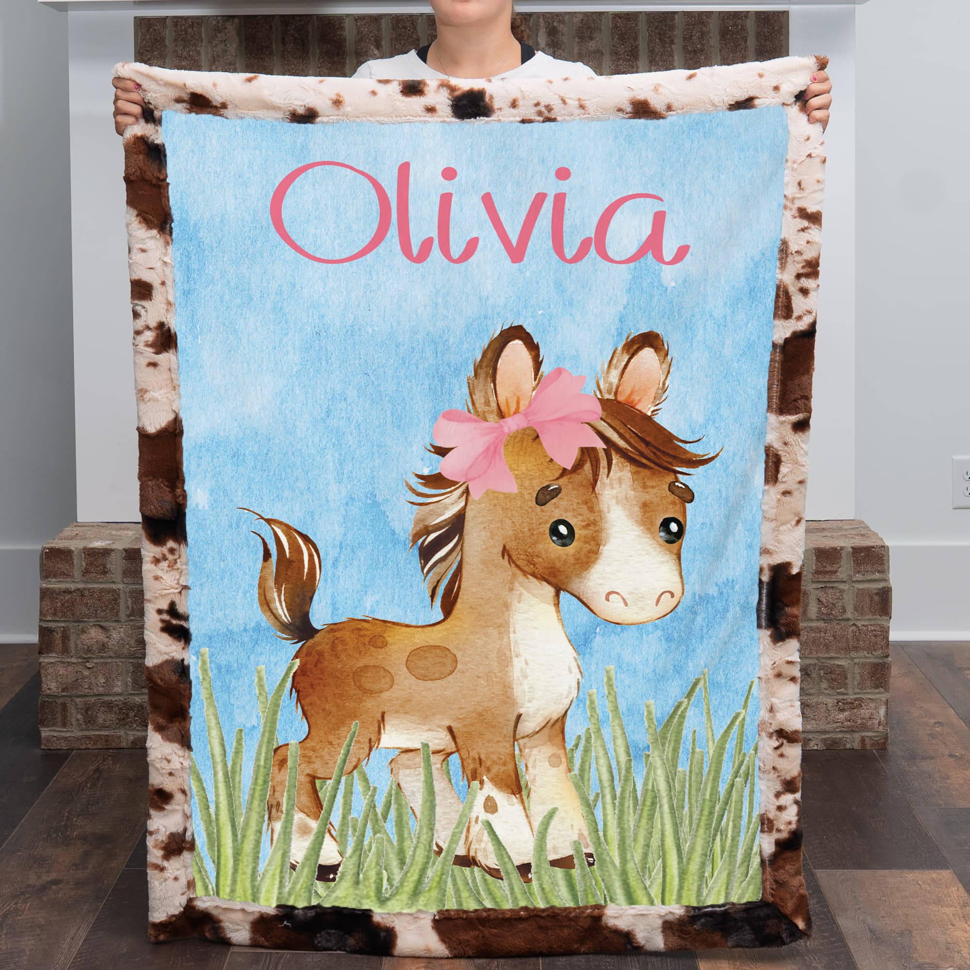 Personalized western pony minky baby blanket with cowhide border and custom name, held up indoors