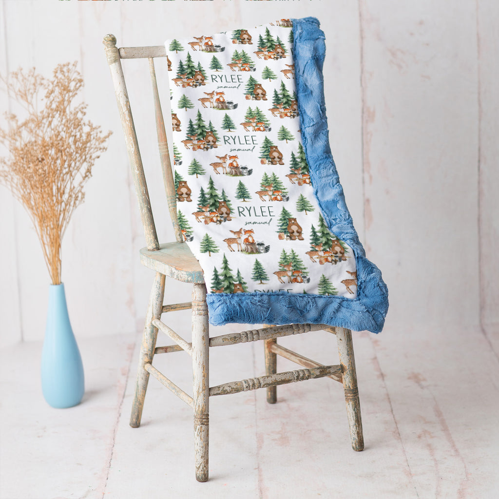 Personalized woodland animal minky blanket with blue trim draped over vintage chair beside dried flowers