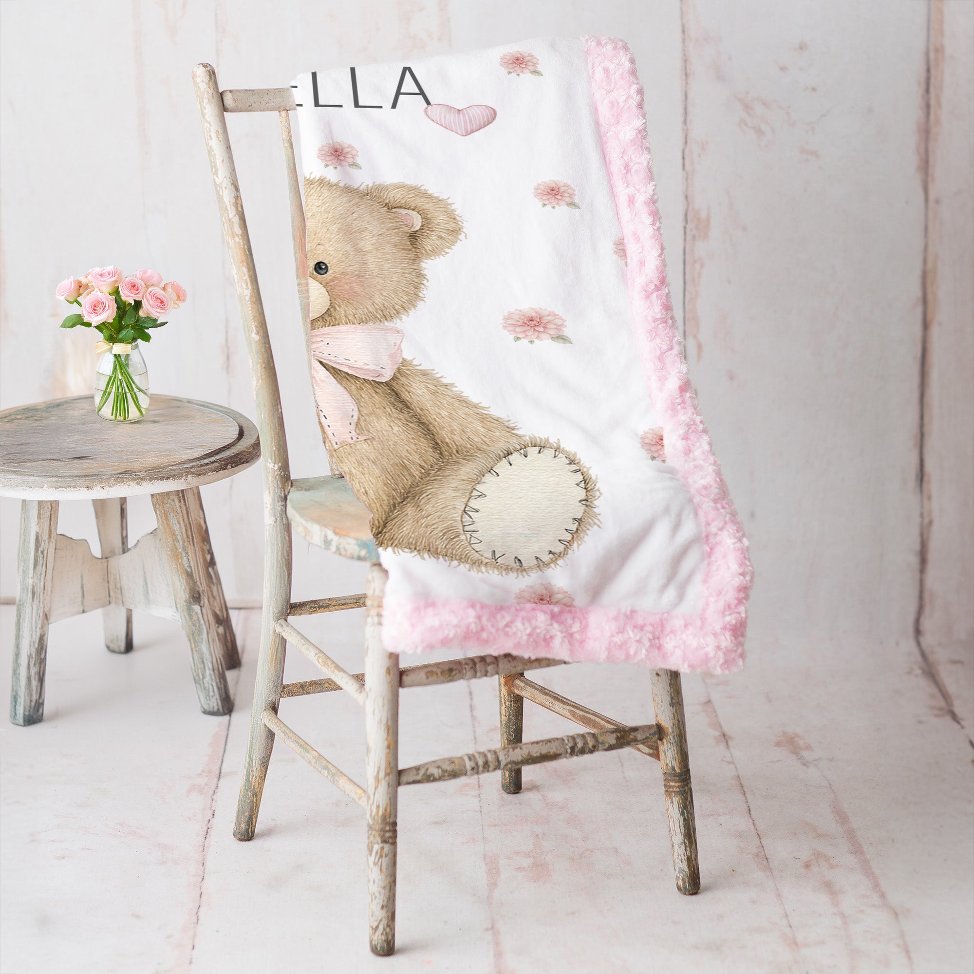 Pink teddy bear floral baby blanket draped over a rustic wooden chair beside a vase of pink roses