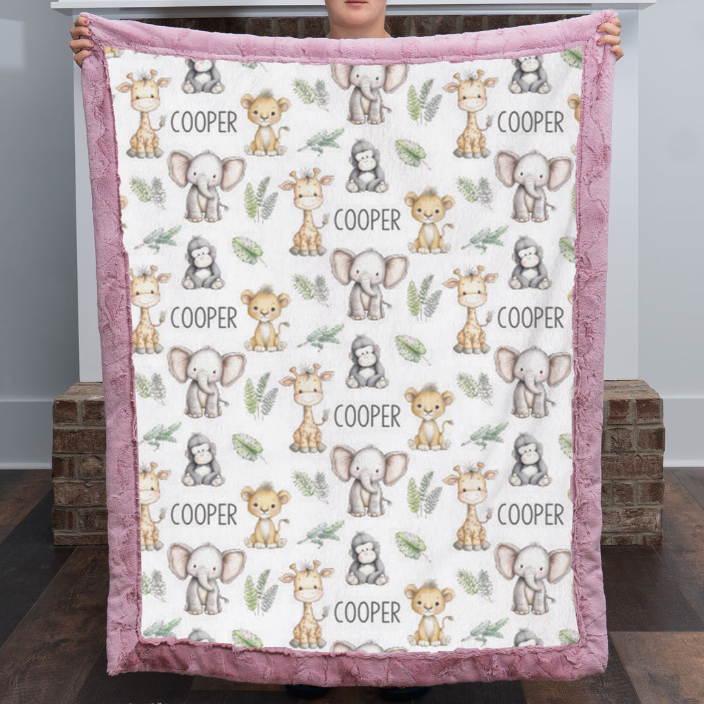 A person holds up a plush minky blanket featuring a repeating pattern of cute safari animals (giraffes, elephants, monkeys, and toddlers) with the name 'COOPER' printed in black text, finished with a dusty pink border.