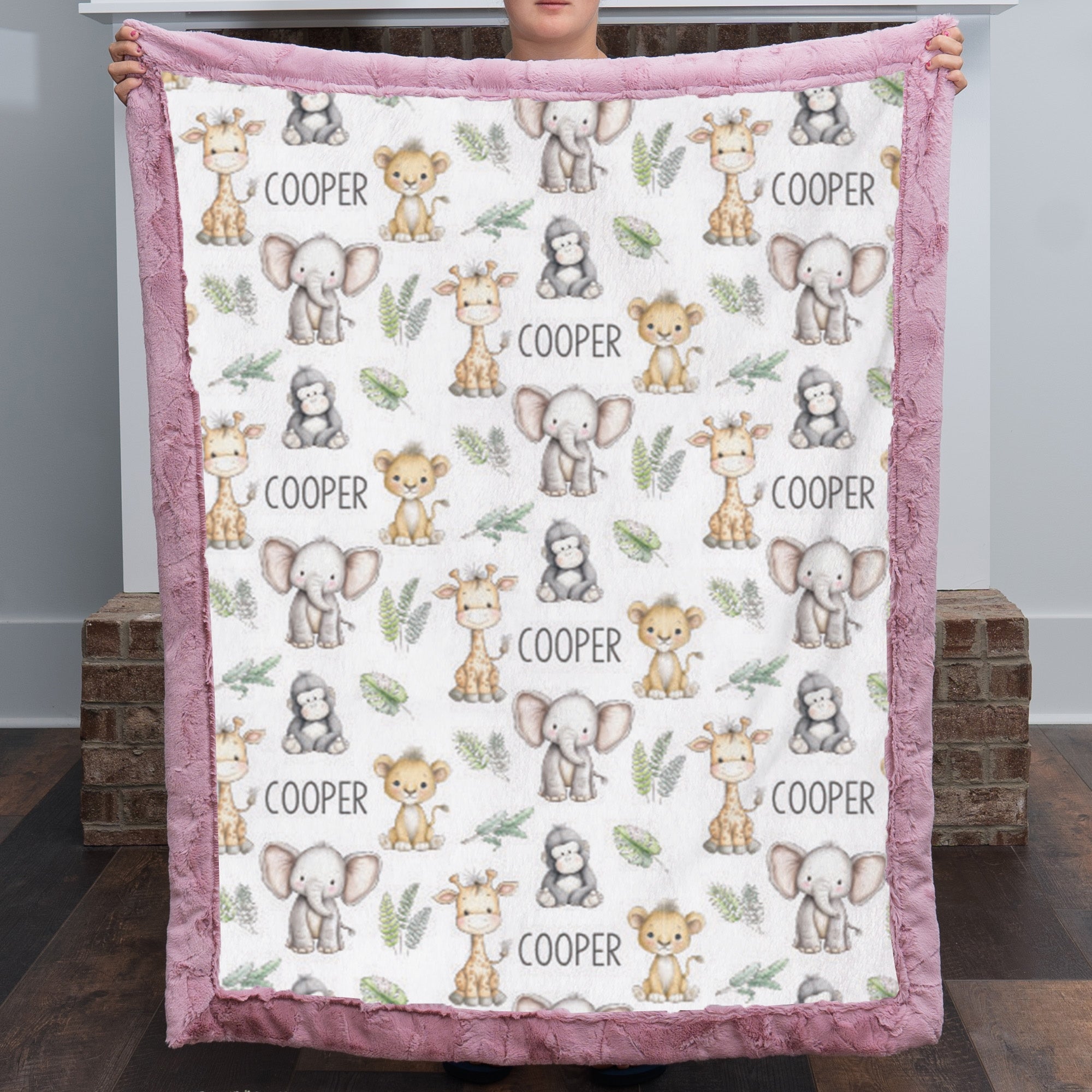 A person holds up a plush minky blanket featuring a repeating pattern of cute safari animals (giraffes, elephants, monkeys, and toddlers) with the name 'COOPER' printed in black text, finished with a dusty pink border.