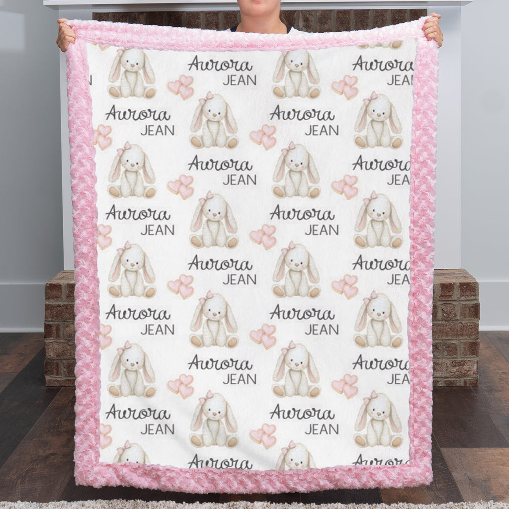 A person holds up a square fleece blanket with a pink fuzzy border. The blanket has a white center printed with a repeating pattern of beige bunny illustrations, pink bows, and the text 'Aurora JEAN'.
