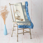 A quilted throw blanket draped over a distressed wooden chair, featuring a vintage-style bunny illustration and blue ruffled borders, set against a soft pink background with a blue vase of dried grass.