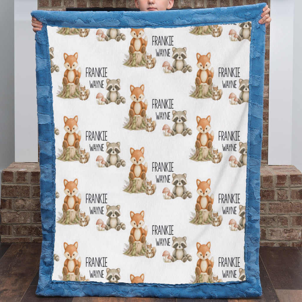 A person holds up a personalized minky quilt blanket with a blue border and a white center printed with cartoon foxes, raccoons, squirrels, and the name 'FRANKIE WAYNE' repeated.