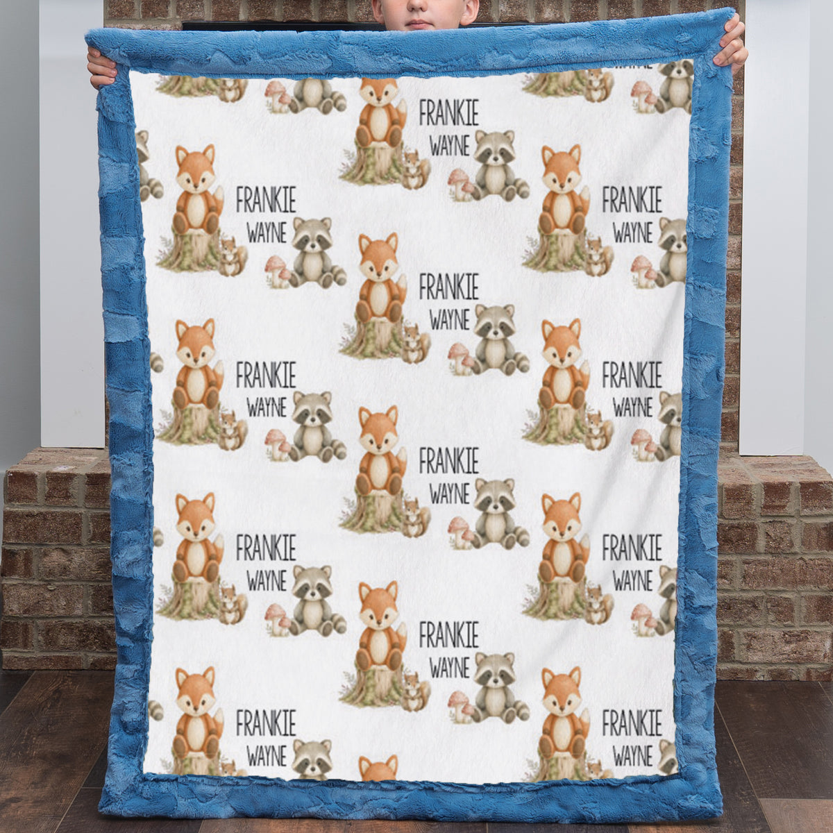 A person holds up a personalized minky quilt blanket with a blue border and a white center printed with cartoon foxes, raccoons, squirrels, and the name 'FRANKIE WAYNE' repeated.