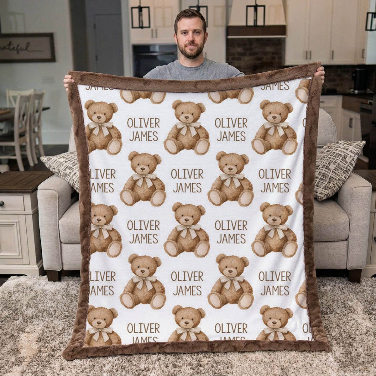 A man stands in a living room holding up a personalized blanket featuring a repeating pattern of teddy bears and the text 'OLIVER JAMES'. The blanket has a white center with brown teddy bears and text, and a thick brown plush border.