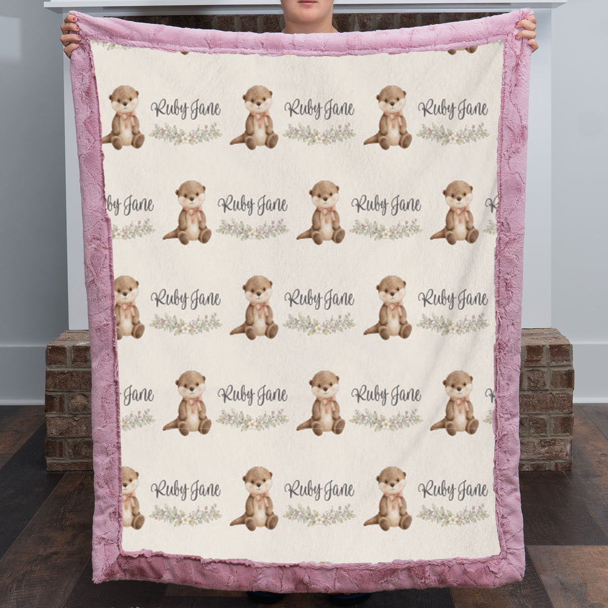 A person holds up a personalized baby blanket featuring a repeating pattern of brown otters and the names 'Ruby Jane' on a cream background, with a pink plush border.