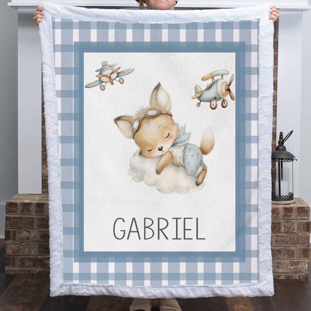 A person holds up a personalized baby blanket featuring a sleeping cartoon fox on a cloud, two vintage airplanes, and the name 'GABRIEL' at the bottom. The blanket has a blue and white gingham border and is shown in front of a brick fireplace.