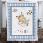 A person holds up a personalized baby blanket featuring a sleeping cartoon fox on a cloud, two vintage airplanes, and the name 'GABRIEL' at the bottom. The blanket has a blue and white gingham border and is shown in front of a brick fireplace.