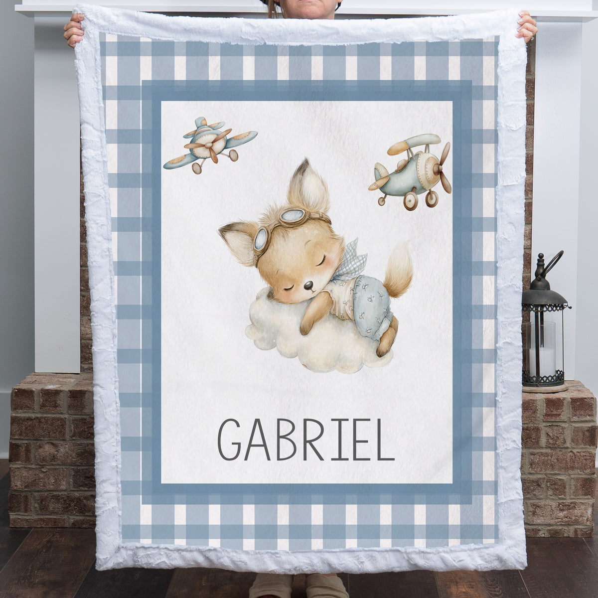 A person holds up a personalized baby blanket featuring a sleeping cartoon fox on a cloud, two vintage airplanes, and the name 'GABRIEL' at the bottom. The blanket has a blue and white gingham border and is shown in front of a brick fireplace.