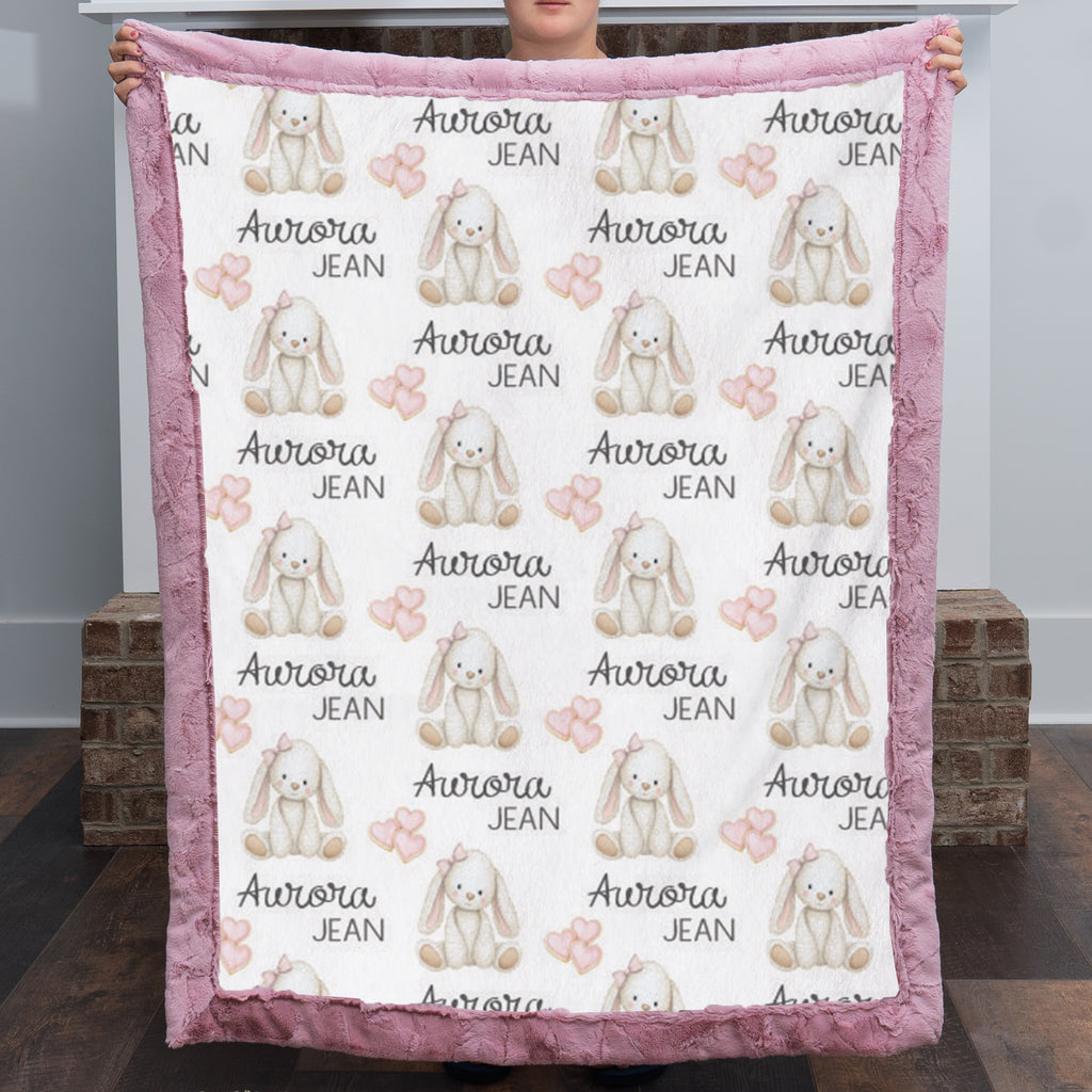 A person holds up a plush blanket featuring a repeating pattern of white rabbits, pink bows, hearts, and the text 'Aurora JEAN'. The blanket has a dusty pink textured border and is photographed in an indoor setting with brick pillars in the background.