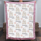 A person holds up a plush blanket featuring a repeating pattern of white rabbits, pink bows, hearts, and the text 'Aurora JEAN'. The blanket has a dusty pink textured border and is photographed in an indoor setting with brick pillars in the background.