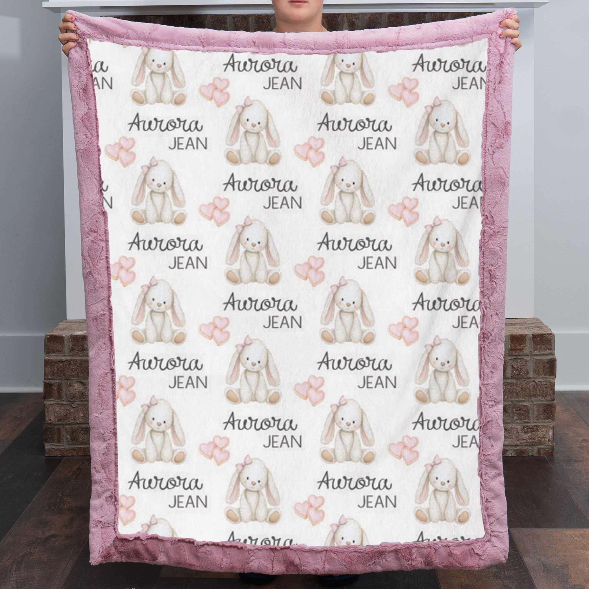 A person holds up a plush blanket featuring a repeating pattern of white rabbits, pink bows, hearts, and the text 'Aurora JEAN'. The blanket has a dusty pink textured border and is photographed in an indoor setting with brick pillars in the background.