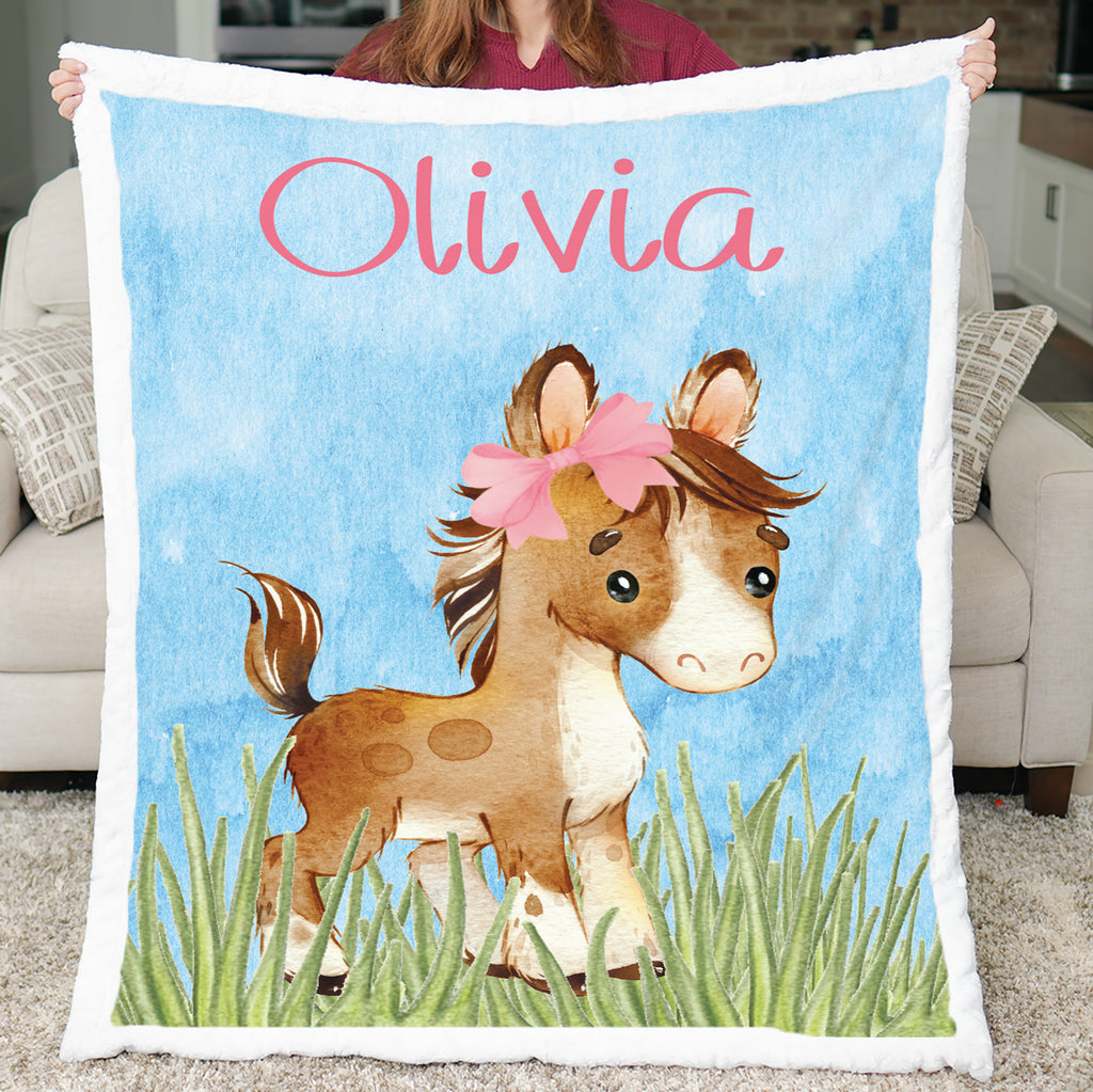 Blanket with a horse illustration and the name 'Olivia' on a blue background, held by a person in a living room.
