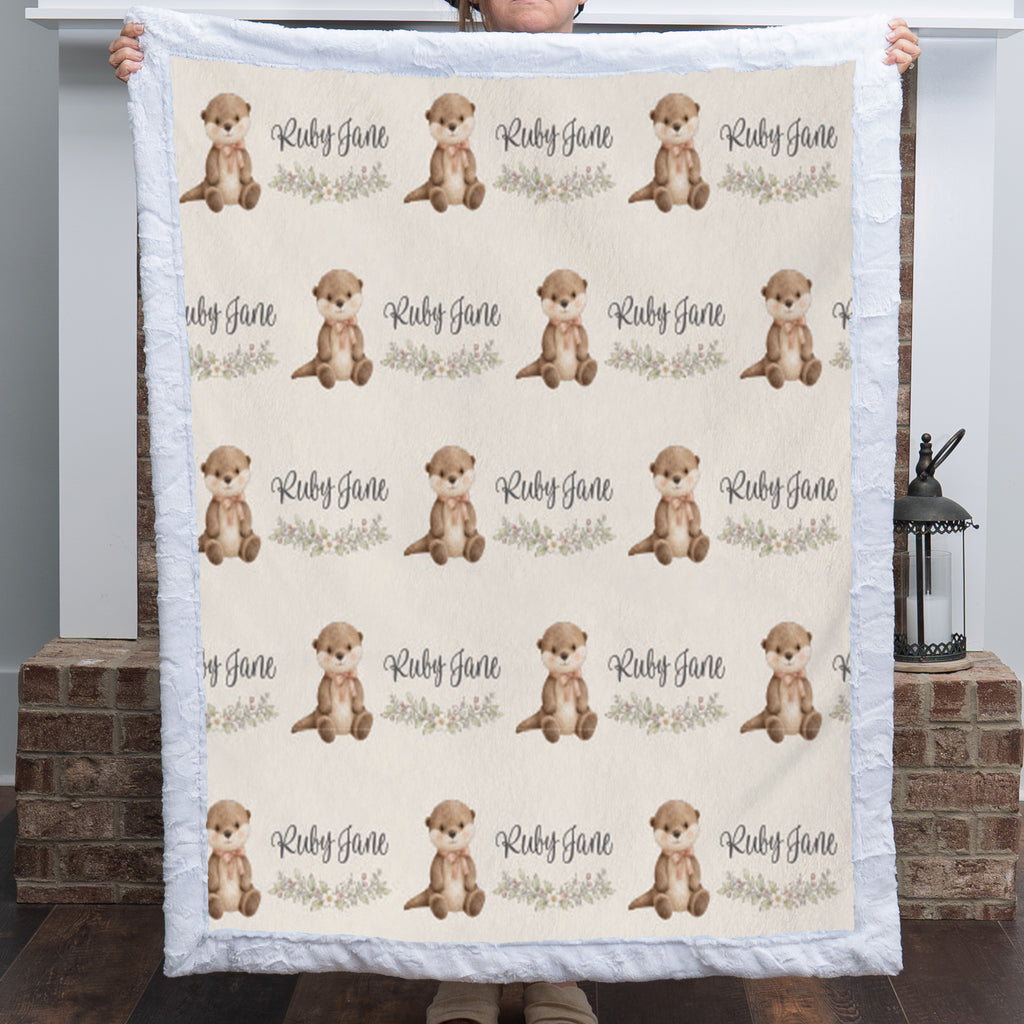 A person holds up a quilted blanket featuring a repeating pattern of brown otters and the text 'Ruby Jane' on a cream background, with a white ruffled border. The background shows a home interior with a brick hearth and a lantern.
