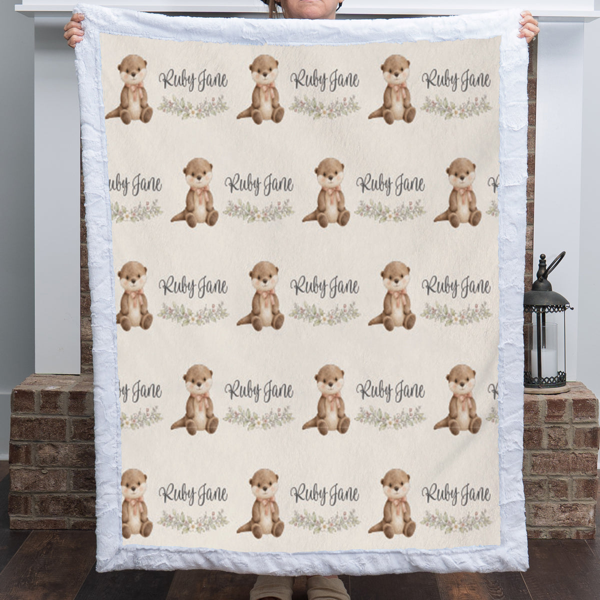 A person holds up a quilted blanket featuring a repeating pattern of brown otters and the text 'Ruby Jane' on a cream background, with a white ruffled border. The background shows a home interior with a brick hearth and a lantern.