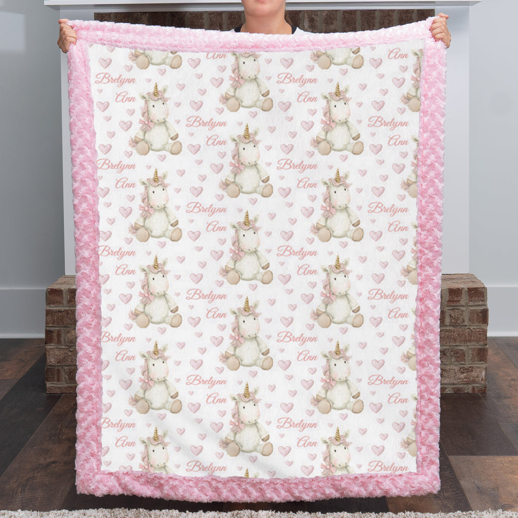 A person holds up a plush fleece blanket featuring a repeating pattern of white and beige unicorns with gold horns, pink hearts, and the words 'Bridleyon Cloe'. The blanket has a thick pink ruffled border and is shown against a home background with wooden floors and brick stools.