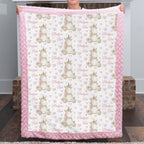 A person holds up a plush fleece blanket featuring a repeating pattern of white and beige unicorns with gold horns, pink hearts, and the words 'Bridleyon Cloe'. The blanket has a thick pink ruffled border and is shown against a home background with wooden floors and brick stools.