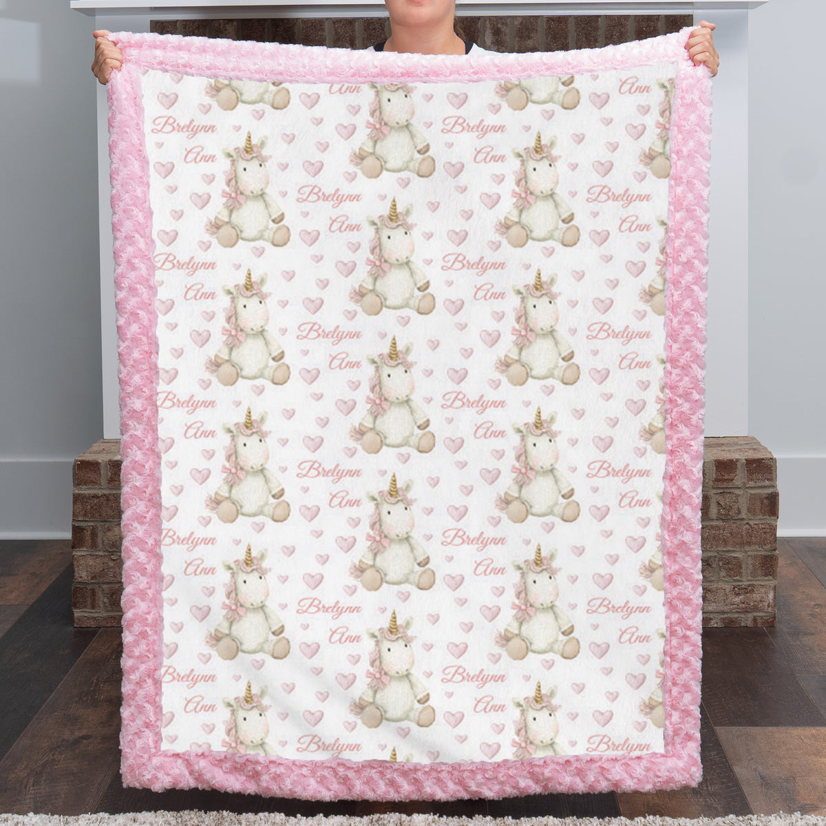 A person holds up a plush fleece blanket featuring a repeating pattern of white and beige unicorns with gold horns, pink hearts, and the words 'Bridleyon Cloe'. The blanket has a thick pink ruffled border and is shown against a home background with wooden floors and brick stools.